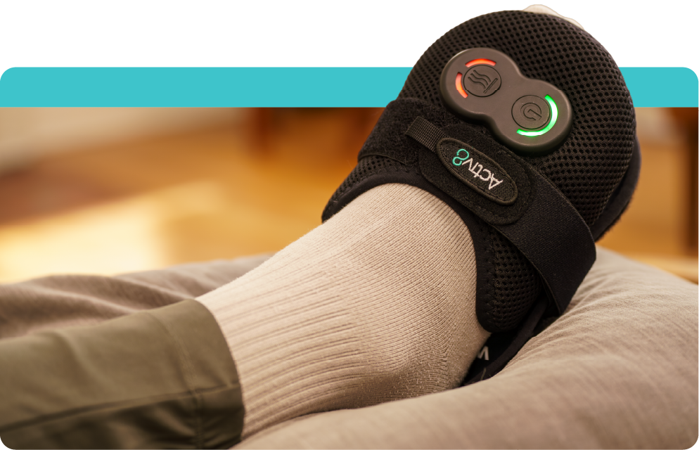 A person's foot resting on a pillow while wearing an Activ8 black compression and heat therapy wrap with glowing green and red power indicators.