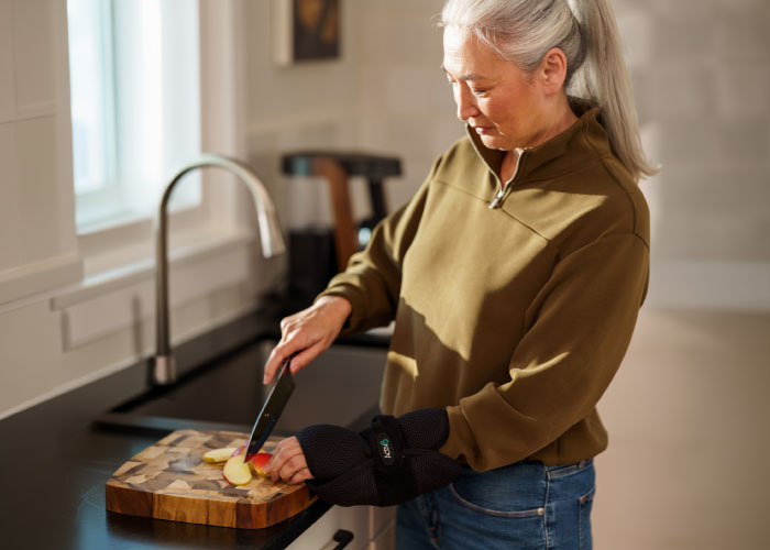 A woman with gray hair wearing a black Activ8 brace on her left wrist while cutting a red apple on a wooden board.